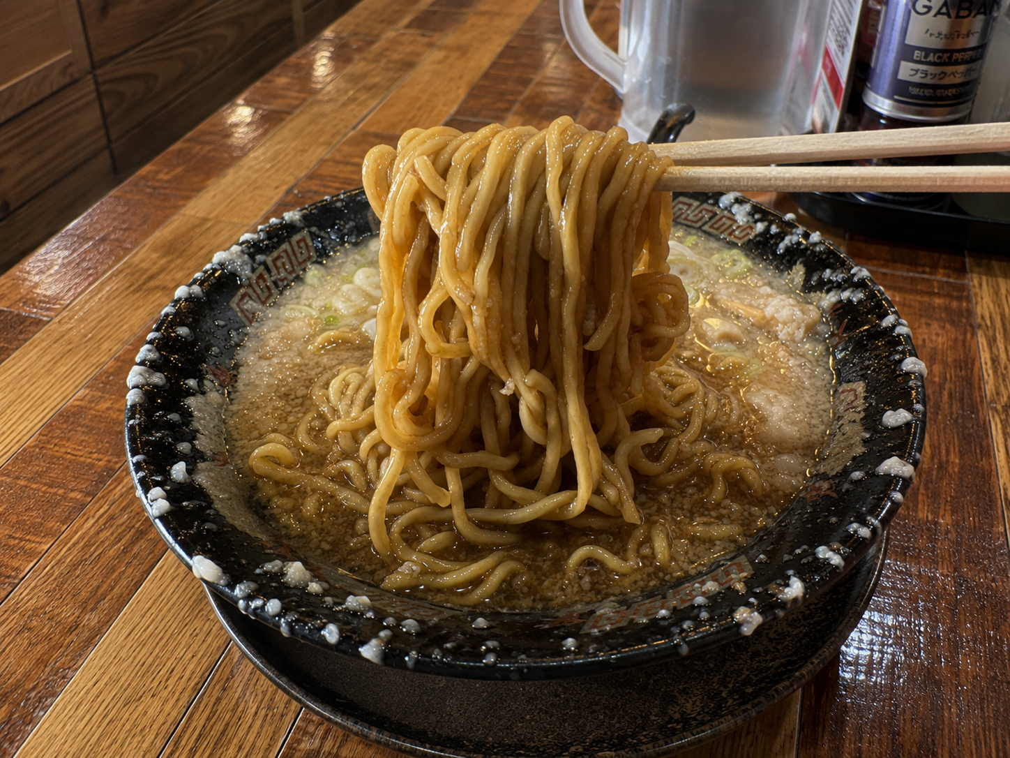 同じ黒い丼から、太めの縮れ麺を箸で持ち上げているところ。背脂の浮いたスープが絡んでいる