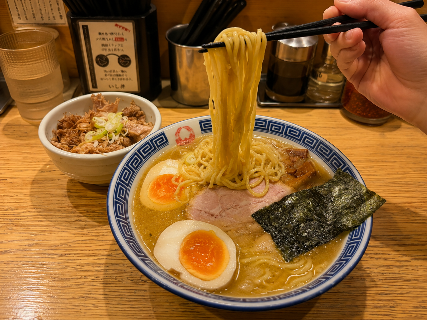 ラーメンの麺を箸で持ち上げているところ。味玉、チャーシュー、海苔が乗り、脇に肉の丼が置かれている