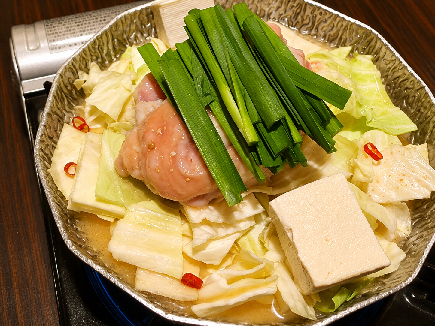 銀色の鍋に鶏肉・キャベツ・豆腐・ニラ・赤唐辛子を入れて煮込んでいる