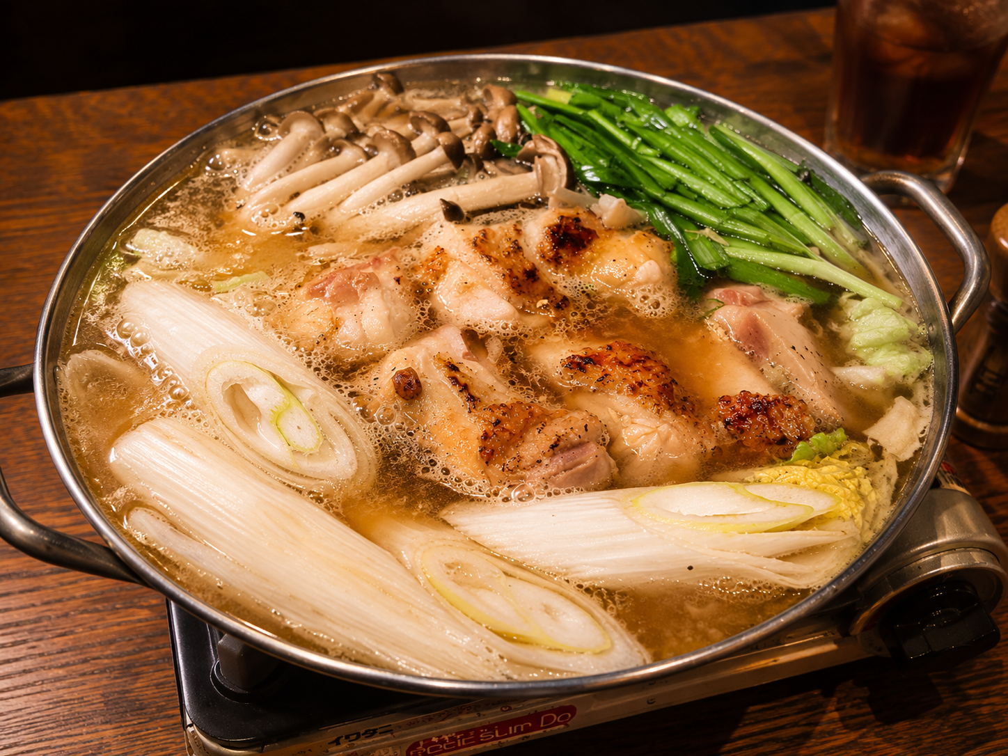 銀色の鍋で煮えている鶏の鍋。長ねぎ、白菜、きのこ、青菜が入っている