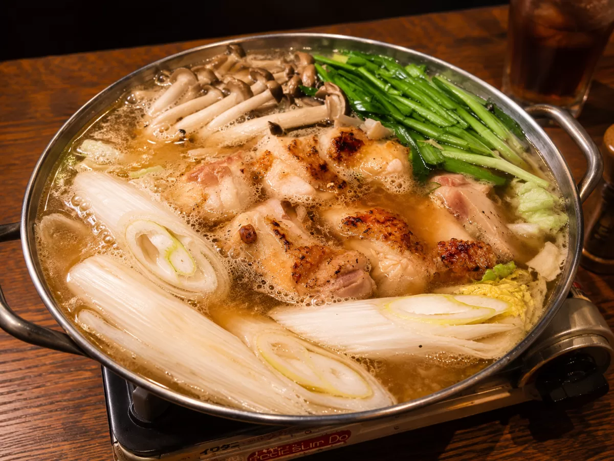 銀色の鍋で煮えている鶏の鍋。長ねぎ、白菜、きのこ、青菜が入っている